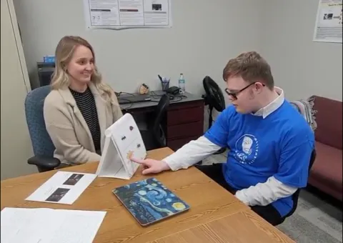 Individual with Down syndrome sitting at table with other adult, pointing to a chart
