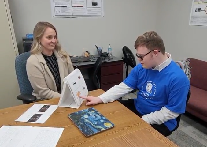 Individual with Down syndrome sitting at table with other adult, pointing to a chart
