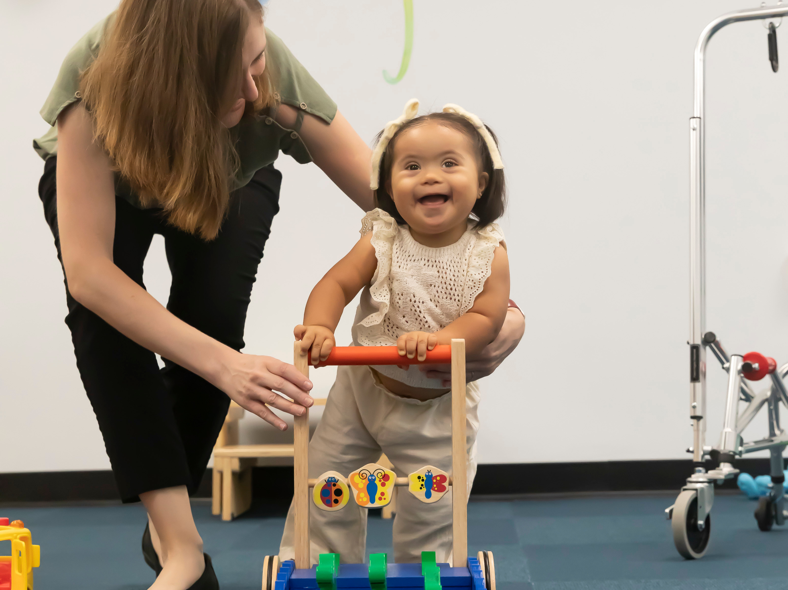 toddler with down syndrome is walking using an assisted mobility device while being supported by an adult